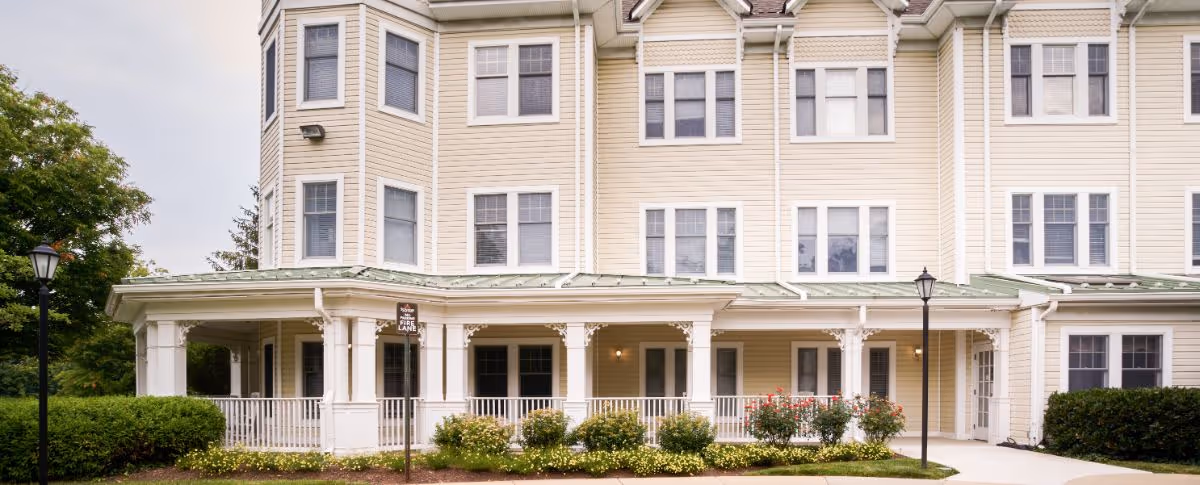 Front exterior of a pale-yellow multi-story senior living building with a covered porch, white railings, and landscaped shrubs.