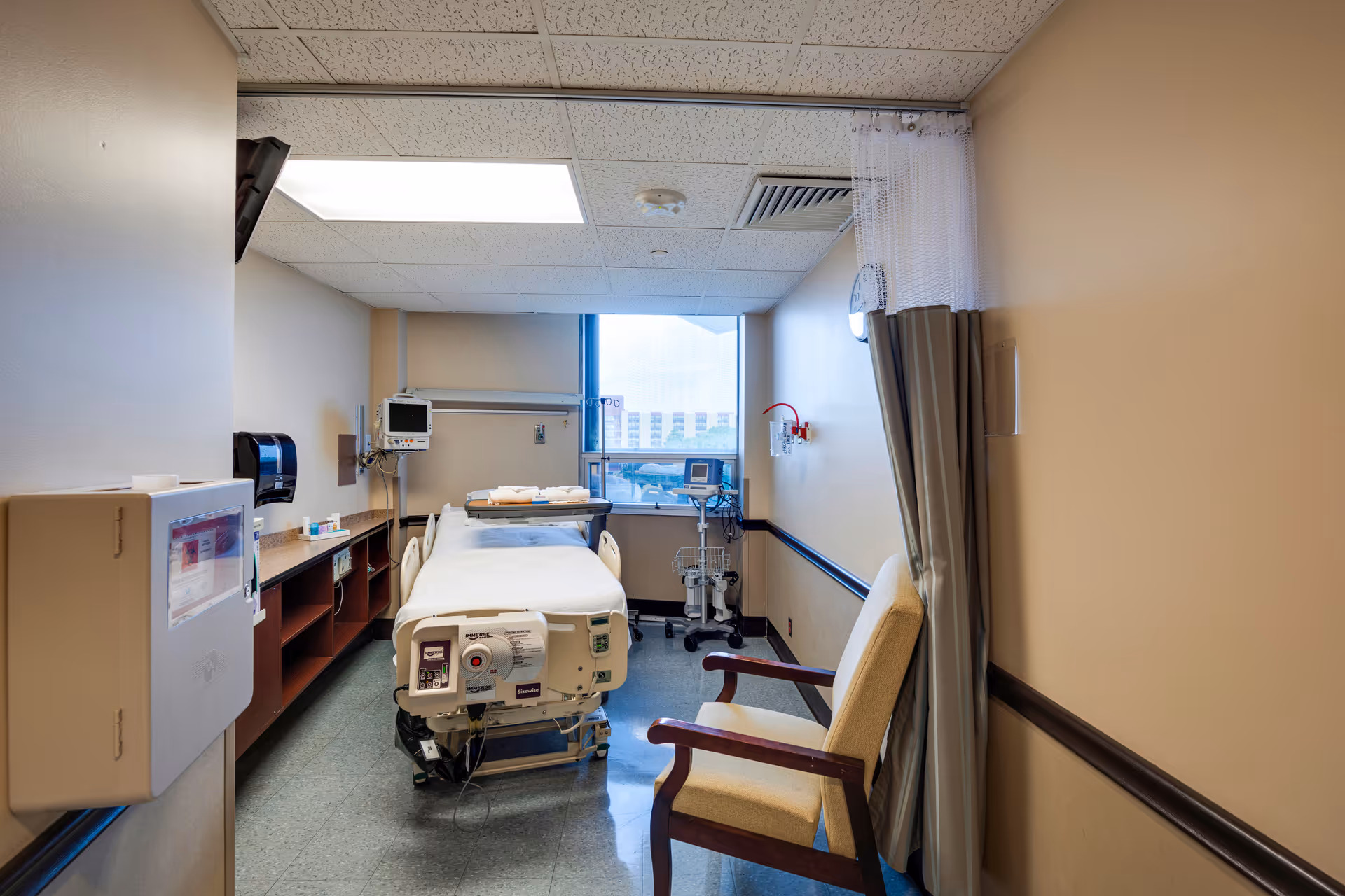 Hospital patient room with bed, medical devices, chair, and privacy curtain.