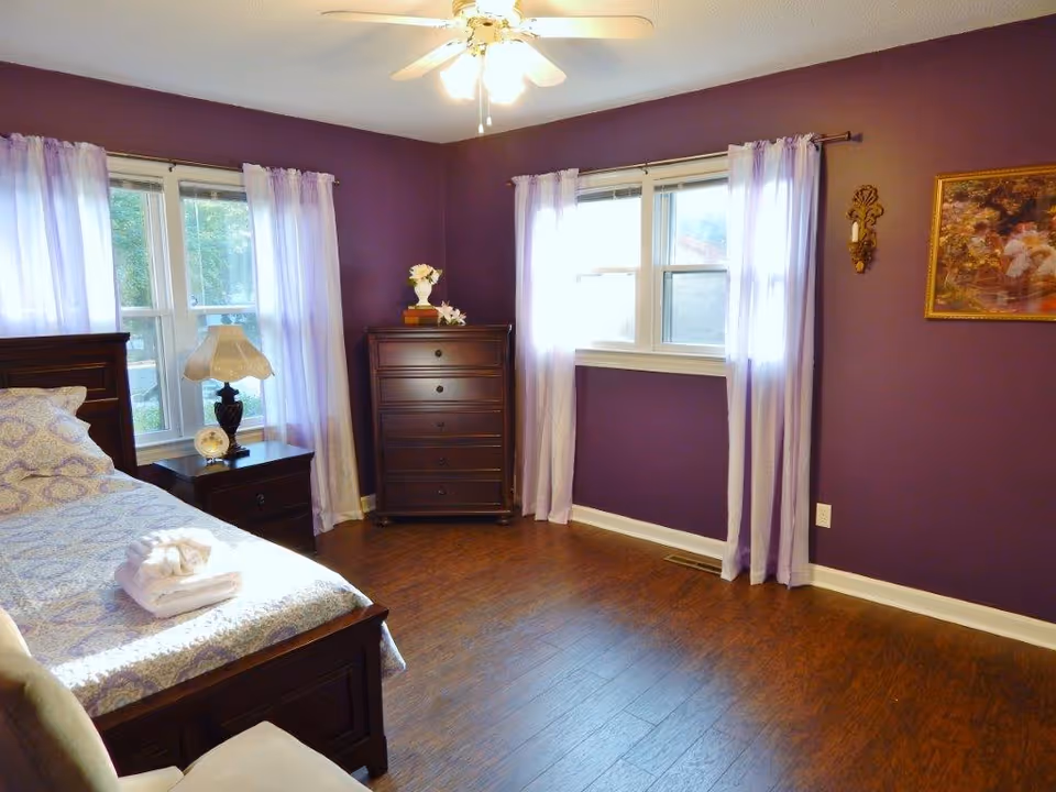Purple-walled bedroom with bed, dresser, nightstand, and sheer lavender curtains.