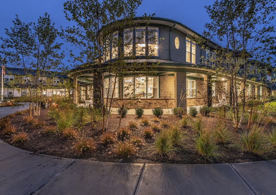 Exterior view of a two-story senior living facility building at dusk with illuminated windows, surrounded by landscaped garden beds with small trees and ornamental grasses, and a curved concrete walkway in the foreground.