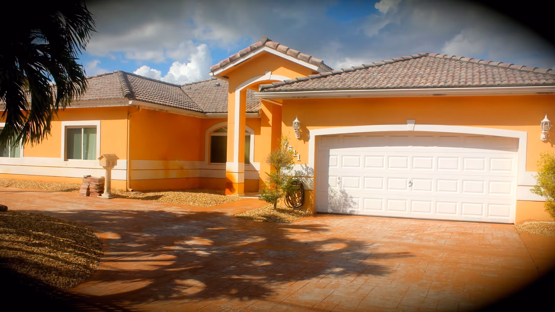 Exterior view of a single-story building with orange walls and a tiled roof, featuring a white garage door and an arched entrance. The driveway is paved and there are small landscaped areas with gravel and plants around the building.