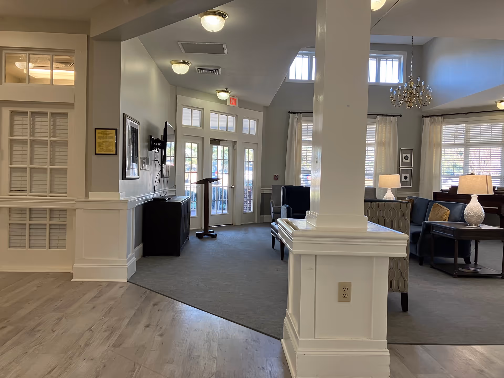Interior view of a senior living facility common area with a mix of carpet and wood flooring. There is a white pillar in the foreground, a seating area with armchairs and sofas, lamps on side tables, a chandelier hanging from the ceiling, and large windows with curtains letting in natural light. A TV is mounted on the wall near the entrance door, which has glass panels and an exit sign above it.