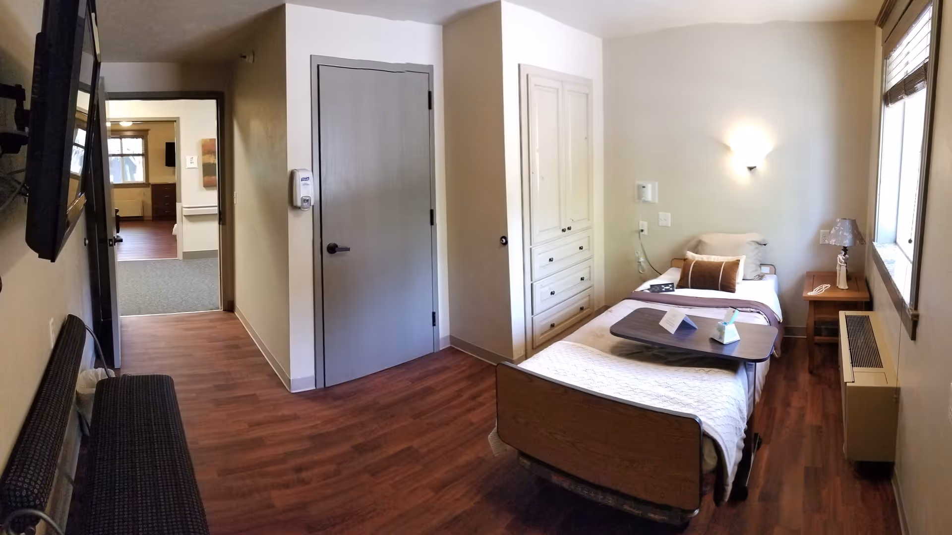 A furnished single-occupancy bedroom in a senior living facility with a bed, dresser, and window.