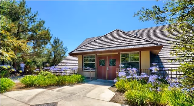 Exterior view of a single-story building with a sloped roof, double wooden doors, and large windows. The entrance is surrounded by green shrubs and purple flowers, with a clear blue sky above.