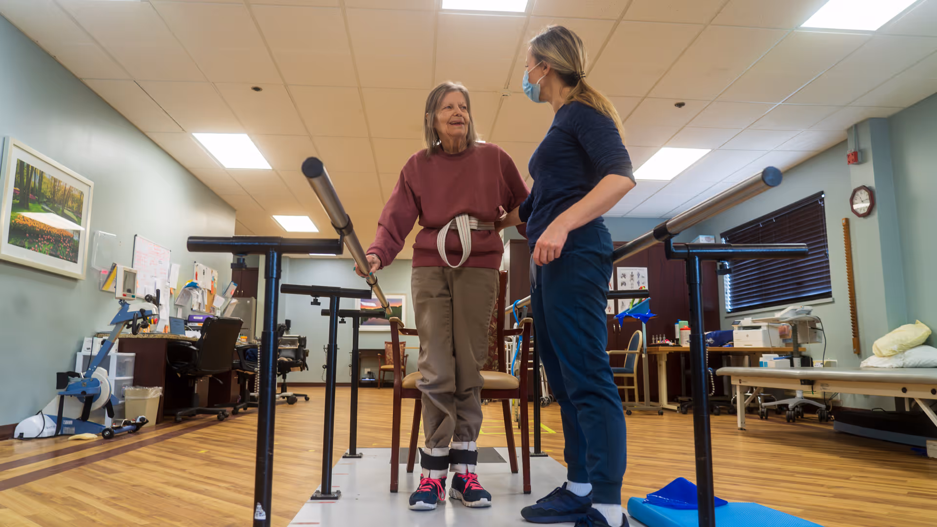 An elderly woman wearing a maroon sweatshirt and beige pants is using parallel bars for walking assistance in a rehabilitation or physical therapy room. A healthcare professional wearing a mask and navy blue scrubs is standing beside her, providing support and guidance. The room has wooden floors, exercise equipment, chairs, and medical supplies visible in the background.
