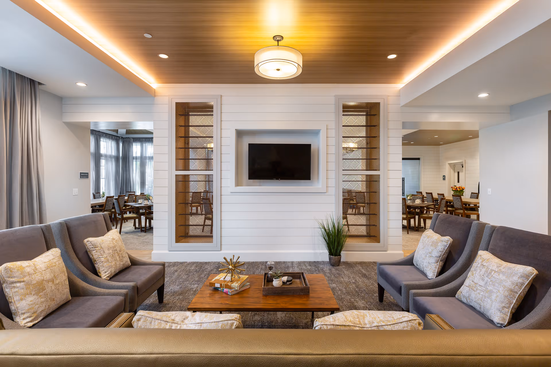 A cozy living room area with four gray armchairs with patterned cushions arranged around a wooden coffee table. The table has decorative items including books and a small plant. The wall opposite the seating area features a mounted flat-screen TV set within a white shiplap wall with two tall, narrow shelves with mesh fronts on either side. The ceiling has wood paneling with a modern light fixture. In the background, there are dining tables and chairs visible through open doorways on either side of the TV wall.