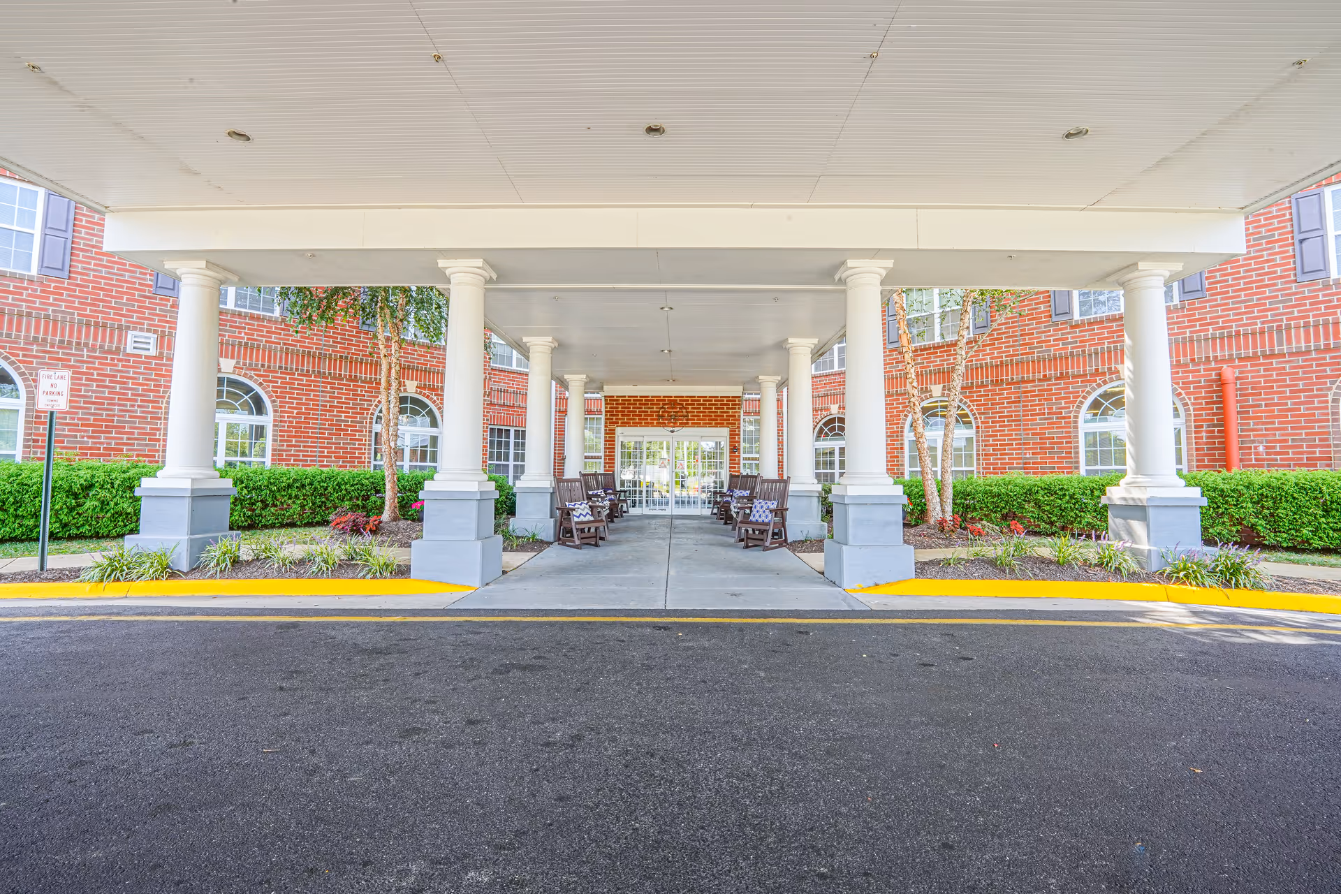 Covered exterior entrance with white columns, benches, and a brick senior living building.
