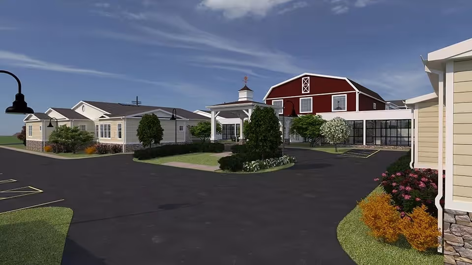 Exterior view of a senior living campus entrance with a driveway, landscaped islands, and cottage-style buildings including a red barn-like main building.