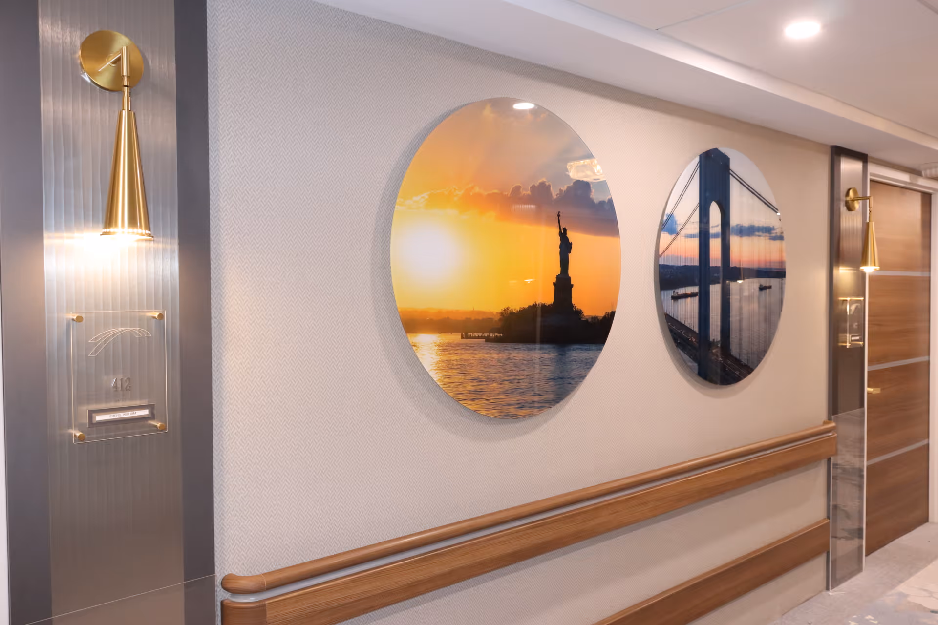 A hallway in Staten Island Care Center with two round wall decorations featuring images of the Statue of Liberty at sunset and a suspension bridge at sunset. The hallway has wooden handrails and modern wall sconces with gold accents. There is a door labeled 412 on the right side.