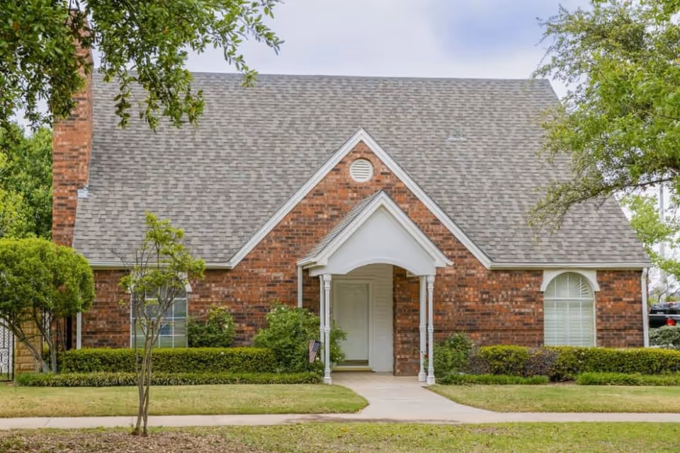 Front exterior of a single-story brick house with a steep shingled roof, central arched entryway, and landscaped lawn.