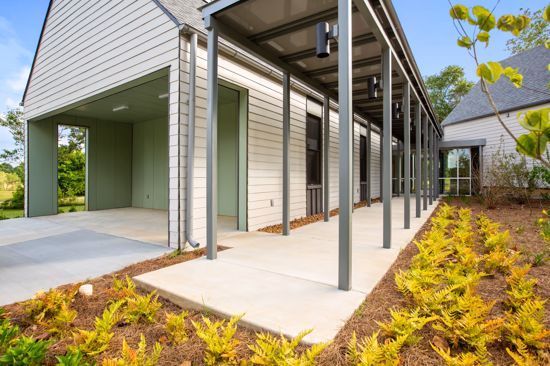 Modern facility exterior with a covered walkway, metal columns, and landscaped plants along the path.