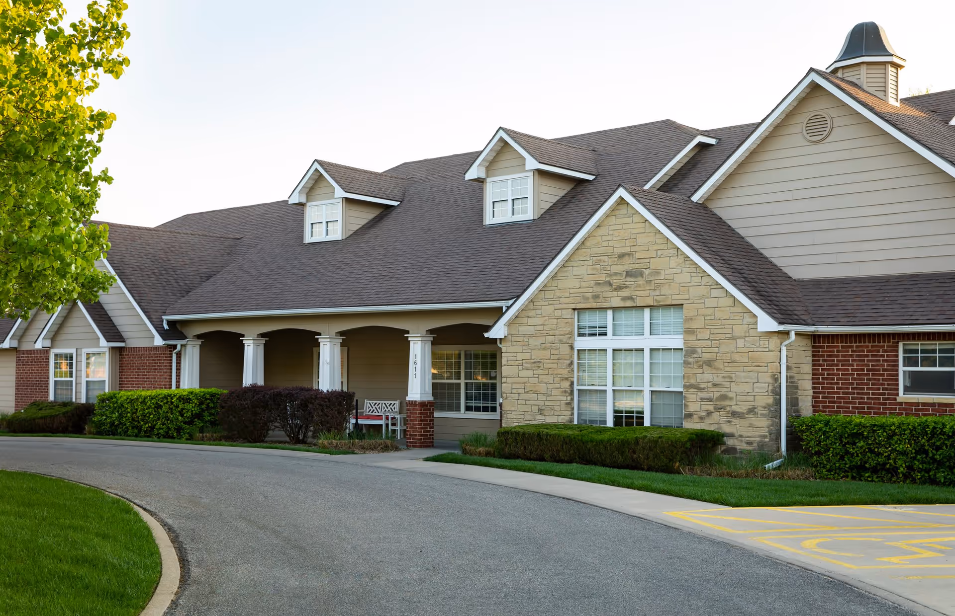 Exterior view of a senior living facility building with a combination of brick, stone, and siding. The building has a covered porch with white columns, several windows, and a curved driveway in front. There is green landscaping including bushes and a tree on the left side.