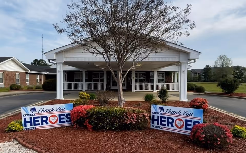 Front exterior view of Liberty Commons Nursing & Rehabilitation Center of Lee County showing a covered entrance with white pillars, a tree in the center of a landscaped area with bushes and flowers, and two signs that read 'Thank You Heroes' placed on either side of the tree.