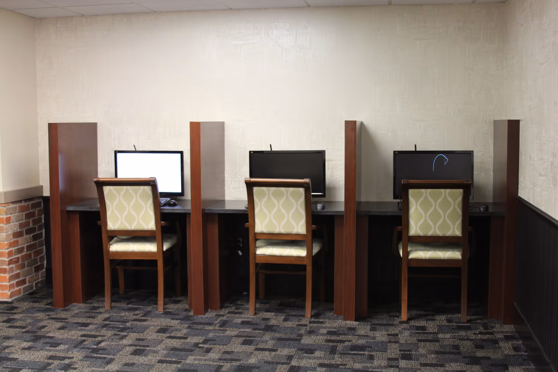 Three computer workstations with chairs separated by wooden dividers against a beige wall.