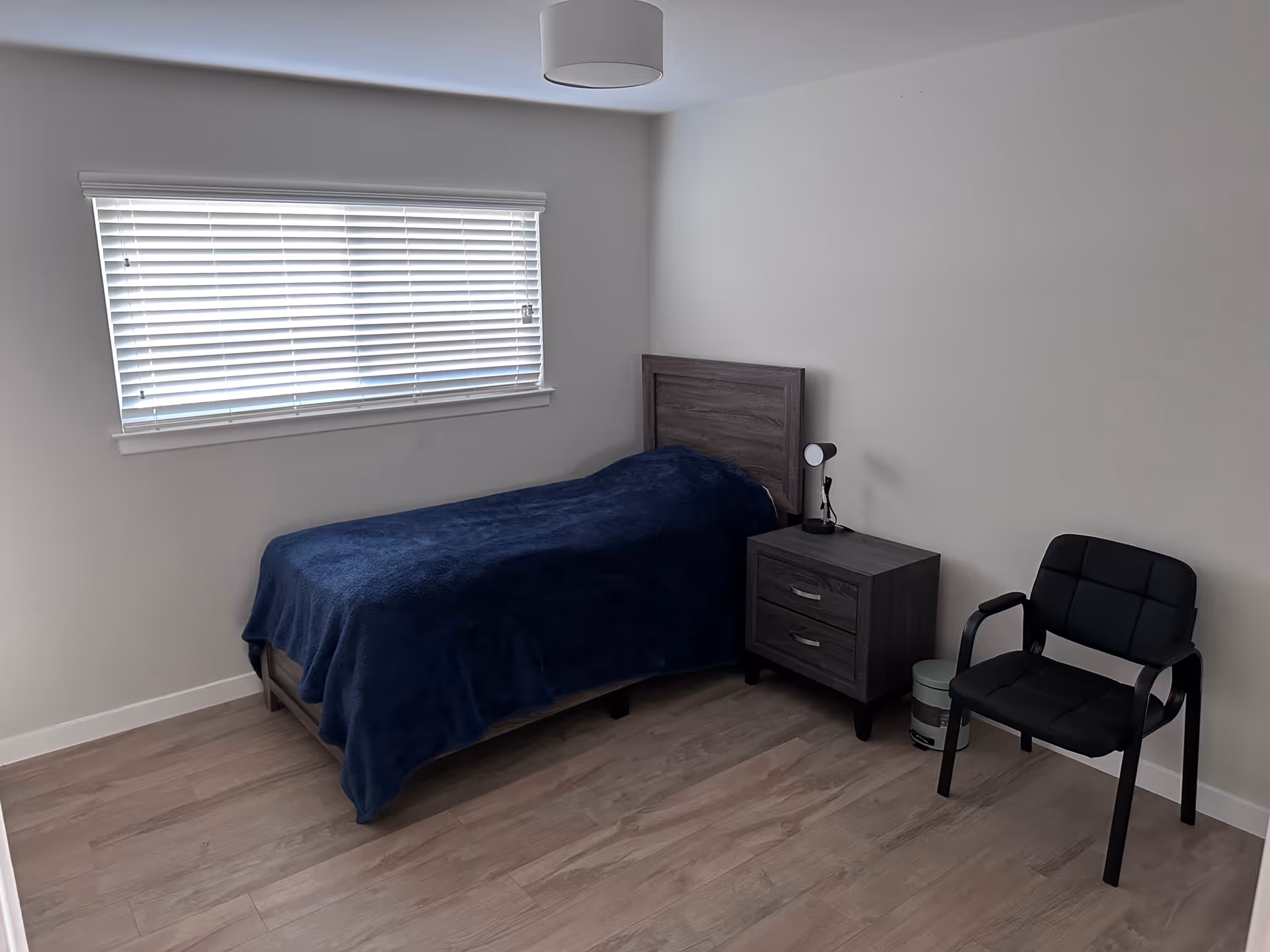 Bedroom with a single bed, nightstand, lamp, and a black chair by the window.