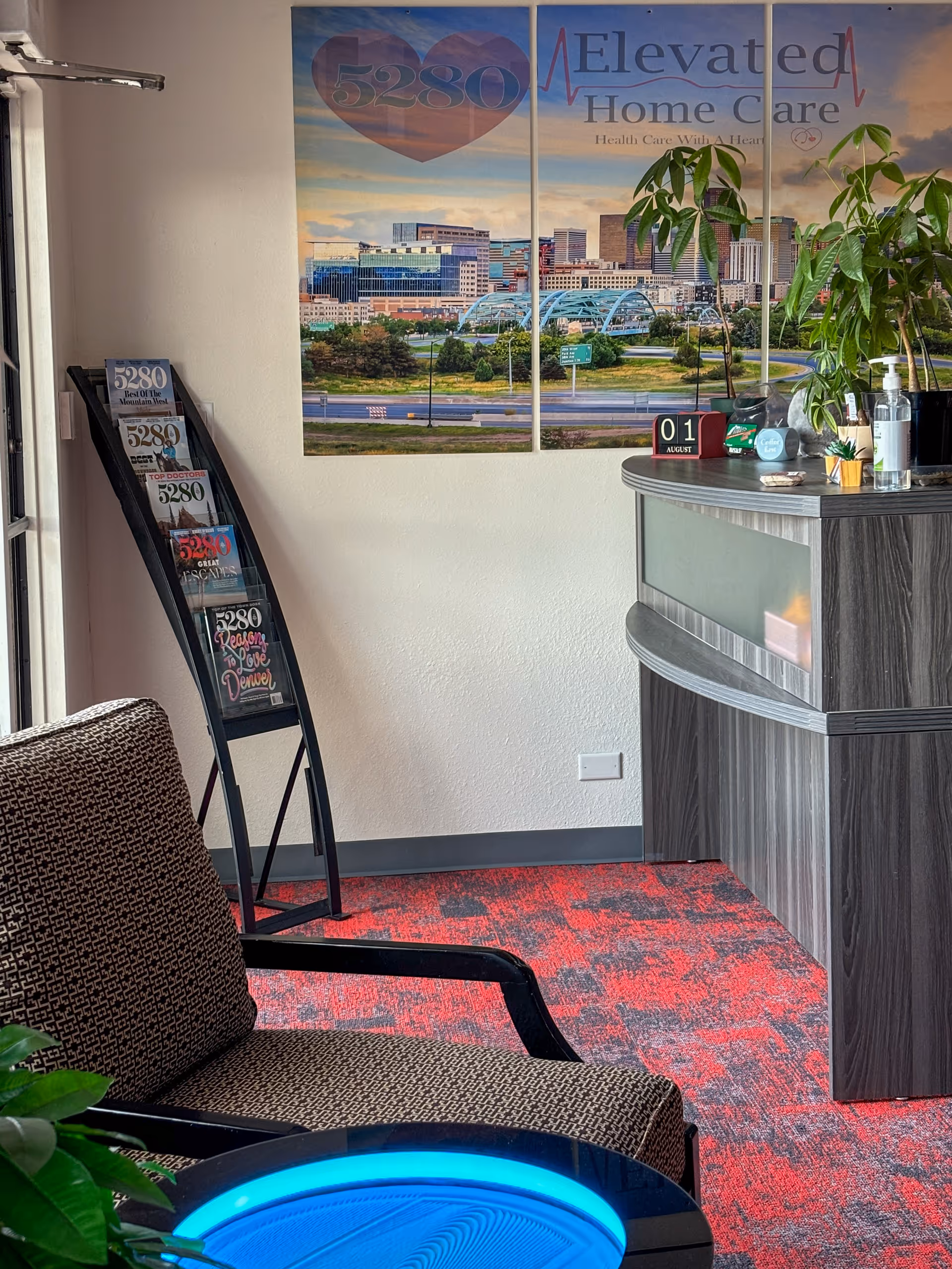 Reception area with curved desk, plant, pamphlet rack, and cityscape poster reading 5280 Elevated Home Care.