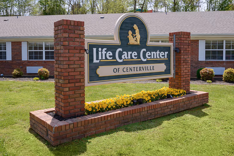A large outdoor sign for Life Care Center of Centerville mounted on two brick pillars with a flower bed of yellow flowers at the base. Behind the sign is a single-story brick building with white window shutters and bushes in front.