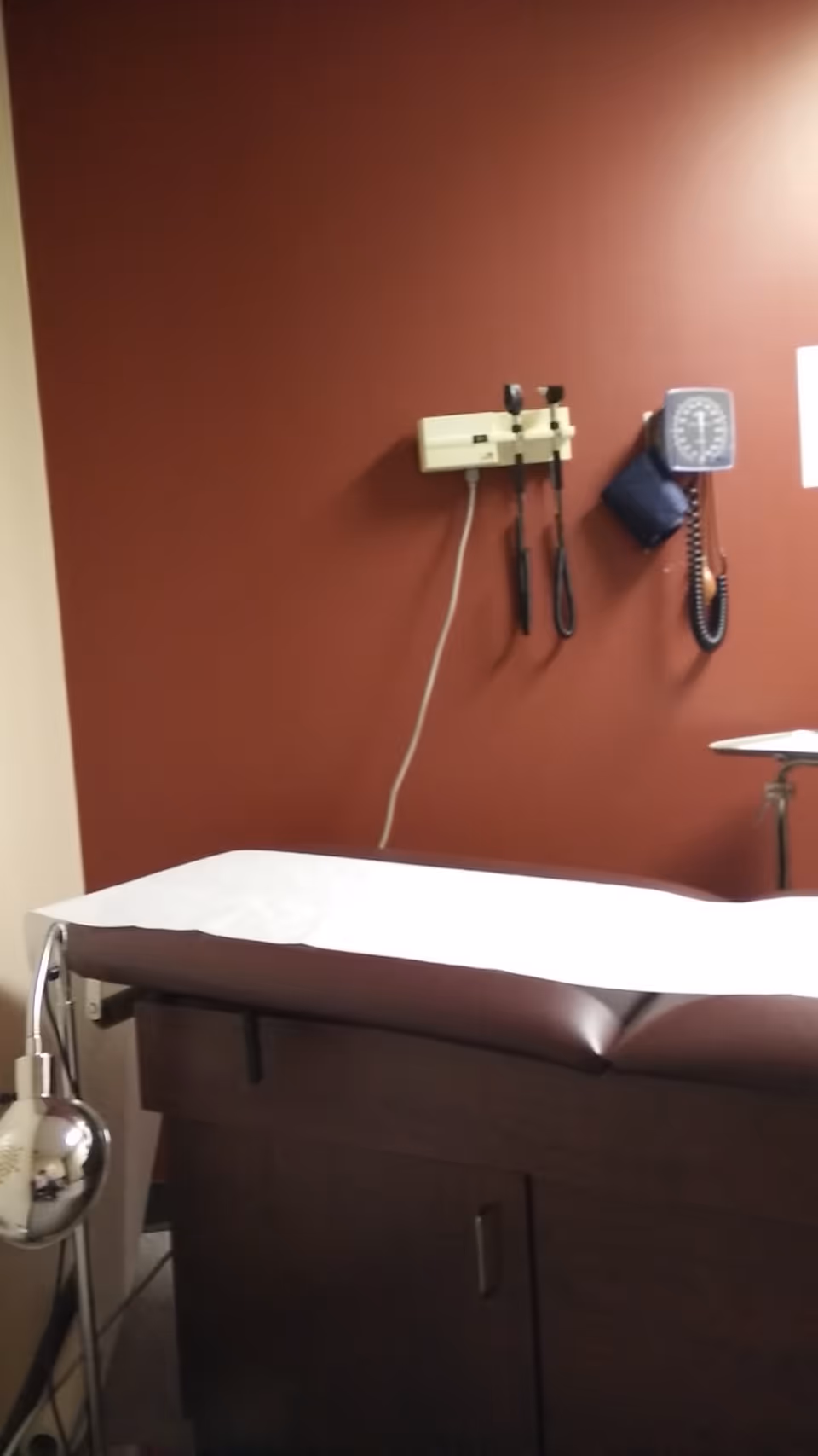 A medical examination table with a blood pressure cuff and stethoscope mounted on a red wall.