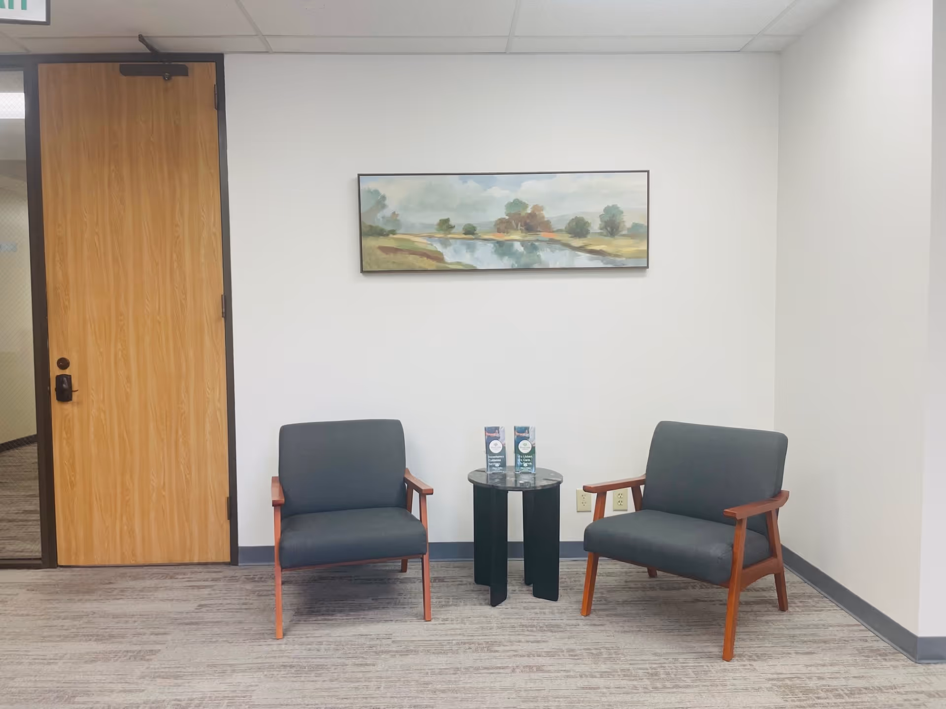 Two gray armchairs face a glass-topped table with brochures in a small lobby area.