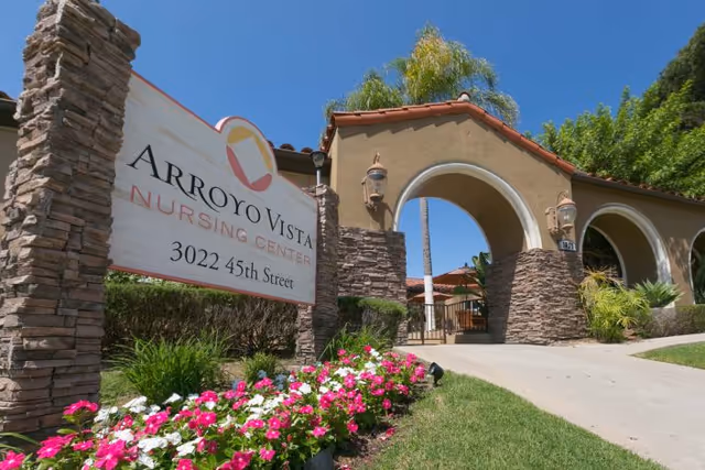 Entrance to Arroyo Vista Nursing Center showing the facility sign, arched stucco entry, and a flowerbed under a clear blue sky.