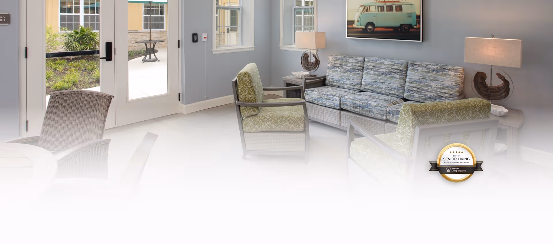 A bright and airy living room area with a patterned sofa, two armchairs, a side table with a lamp, and a framed picture of a vintage van on the wall. Large glass doors open to an outdoor patio with greenery visible outside.