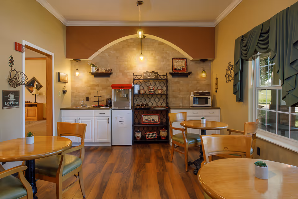 A cozy dining area with wooden tables and chairs, a small kitchenette with white cabinets, a mini fridge, microwave, and a black metal shelf holding decorative items. The room has warm lighting with hanging pendant lights, a large window with green curtains, and wood flooring.