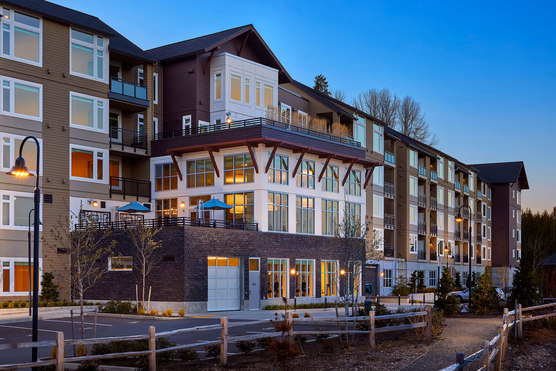 Exterior view of a multi-story senior living facility building at dusk with large windows, balconies, outdoor seating areas with umbrellas, street lamps, and a wooden fence along a pathway.