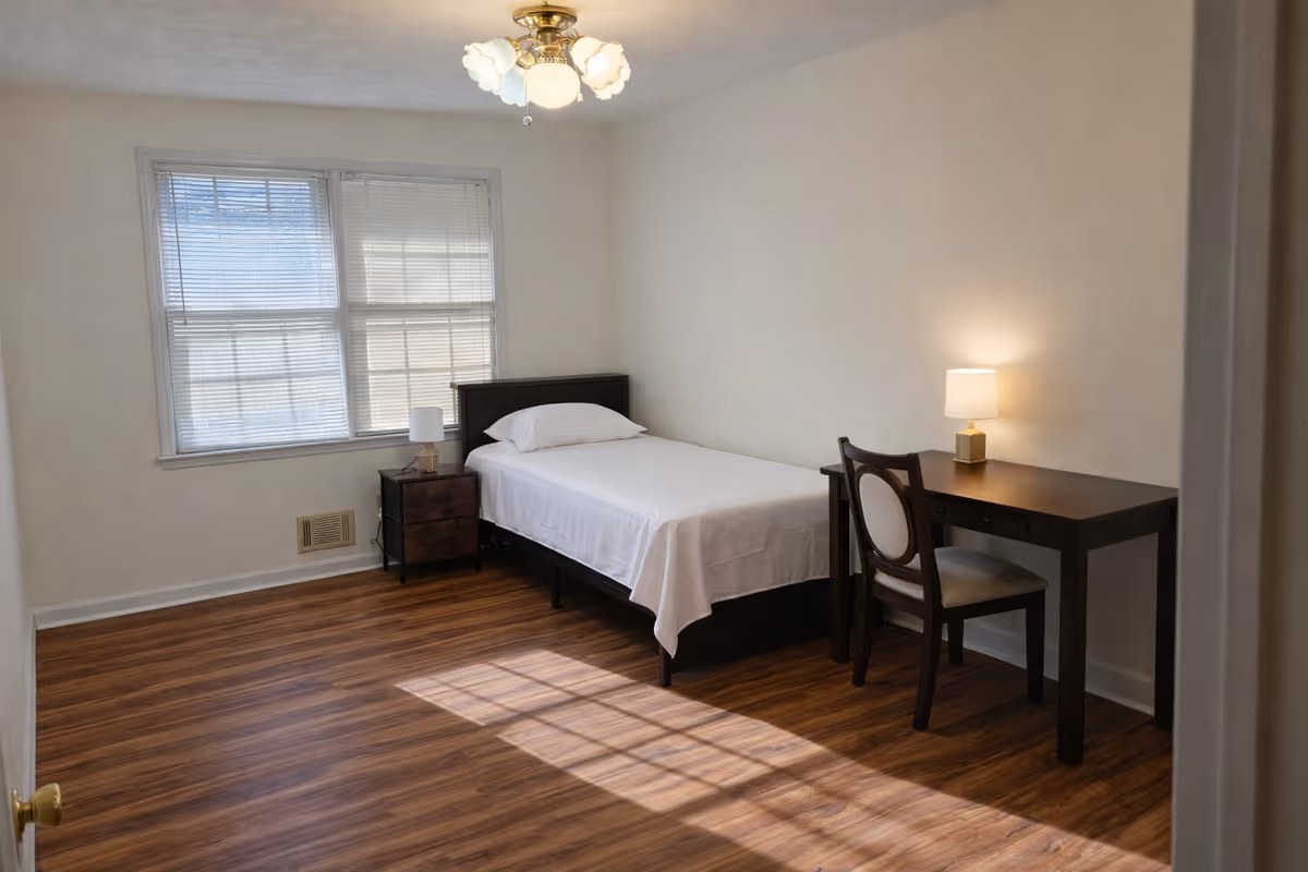 A simple bedroom with a single bed, desk, chair, lamp, and window.