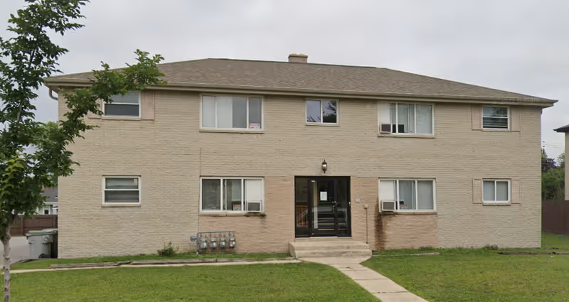 Two-story beige apartment-style senior living building with a central entrance and green lawn.