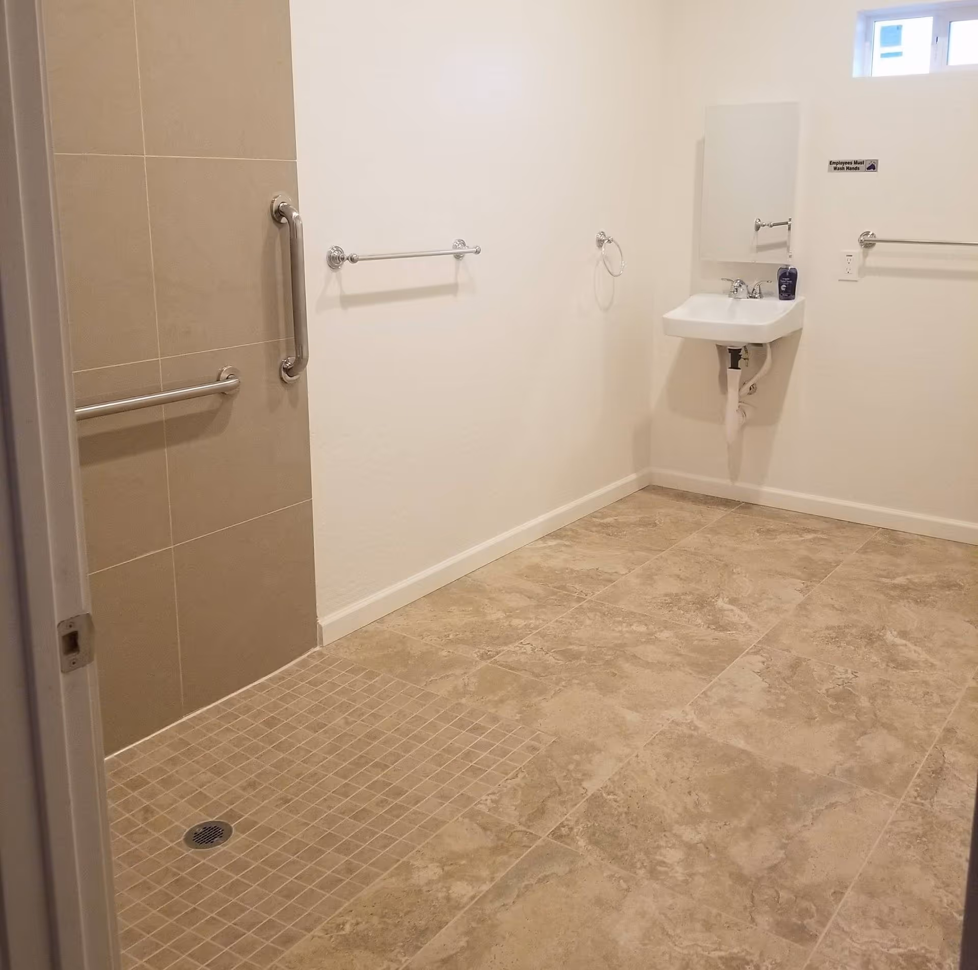 A clean, accessible bathroom with beige tiled floor and shower area. The shower has two metal grab bars on the tiled wall. There is a white wall-mounted sink with a mirror above it, a soap dispenser on the sink, and multiple towel bars on the walls. A small window is visible near the ceiling.