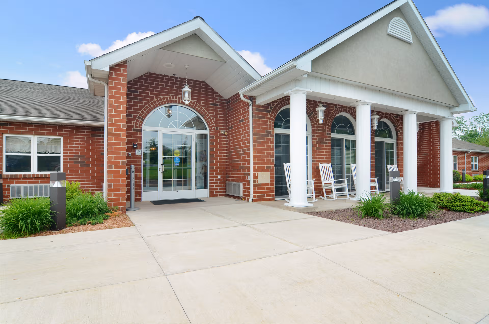 Brick senior living facility entrance with white columns and rocking chairs.