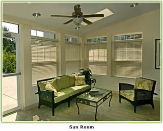 A sunroom with large windows covered by blinds, featuring a green cushioned sofa, a matching armchair, a glass coffee table, a ceiling fan, and a light-colored rug on the floor.