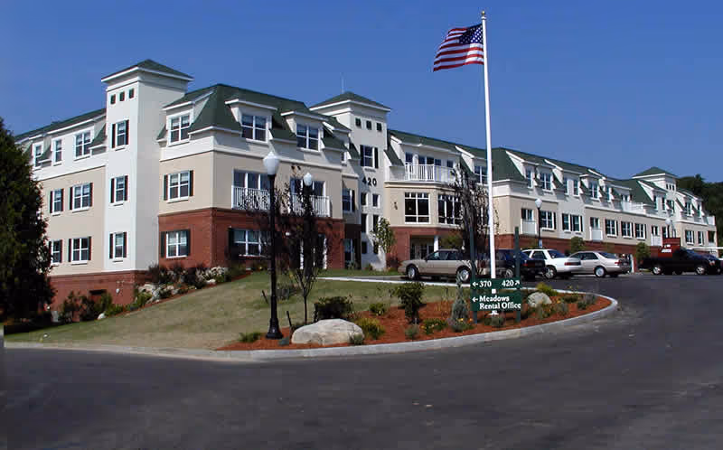 Front exterior view of a multi-story senior living facility with an American flag, landscaped roundabout, and parked cars.