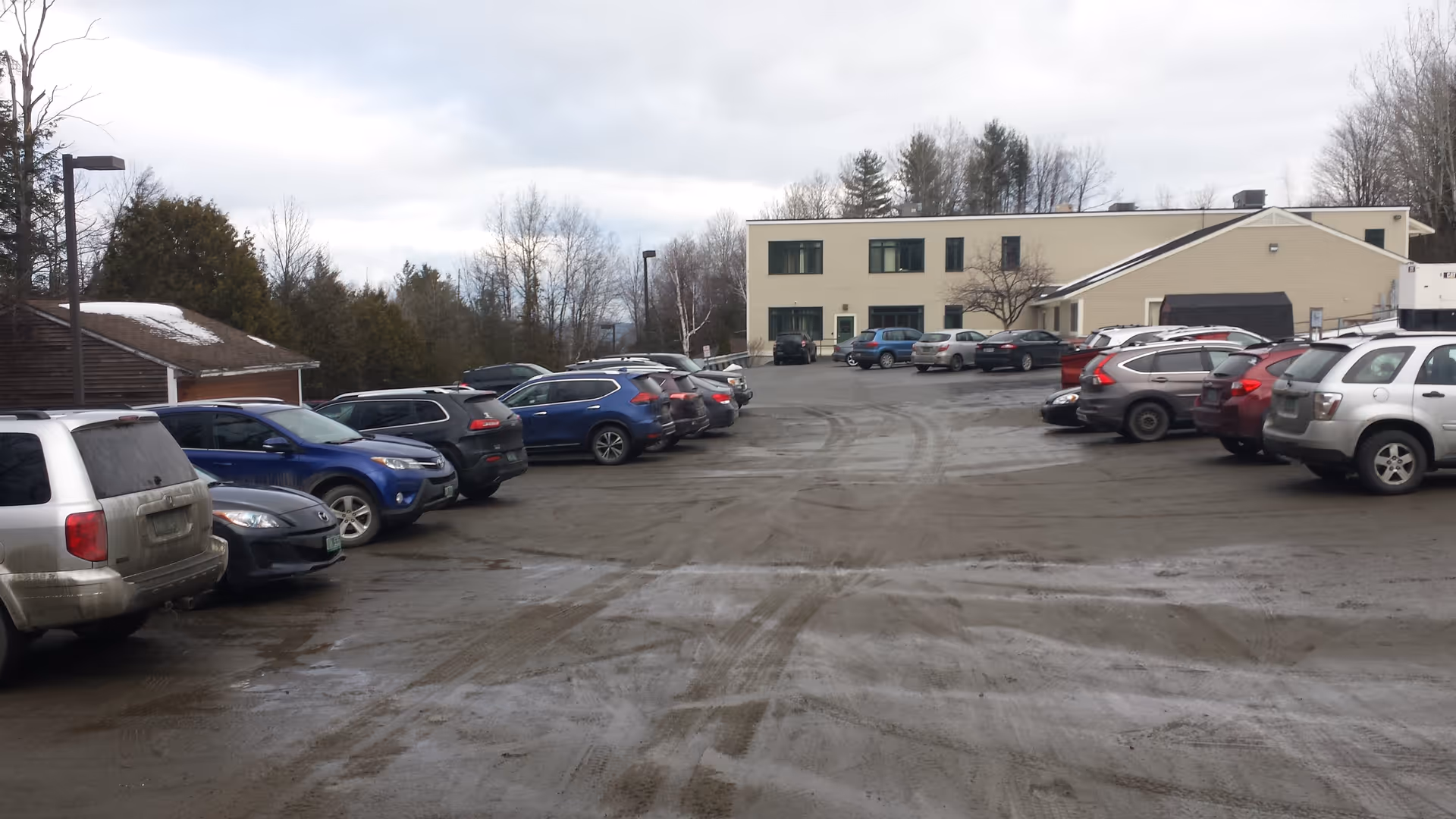 A parking lot full of cars surrounds a beige two-story building under an overcast sky.