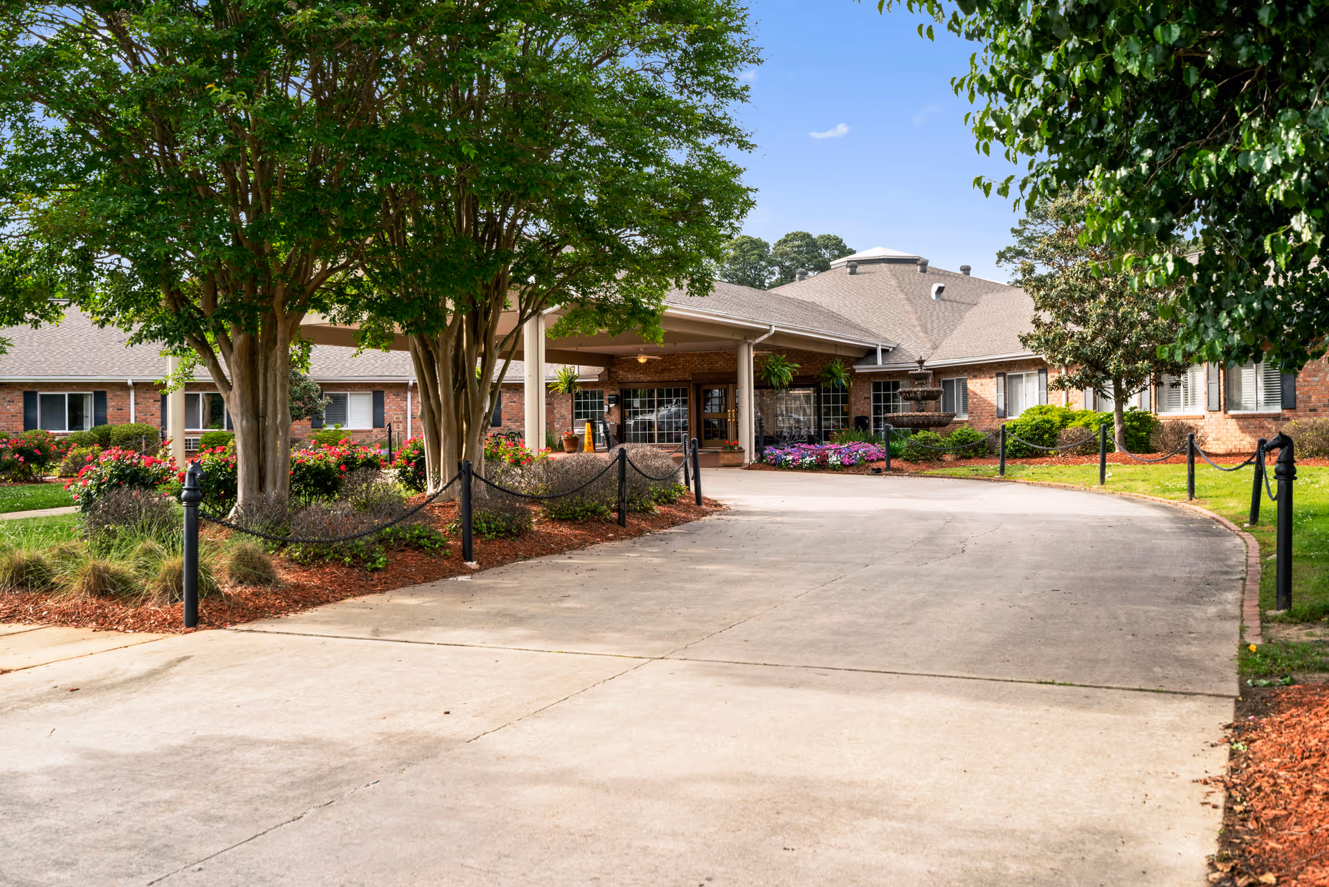 Driveway leading to the entrance of a single-story brick building with a covered porch, surrounded by landscaped gardens with trees, shrubs, and colorful flowers under a clear blue sky.