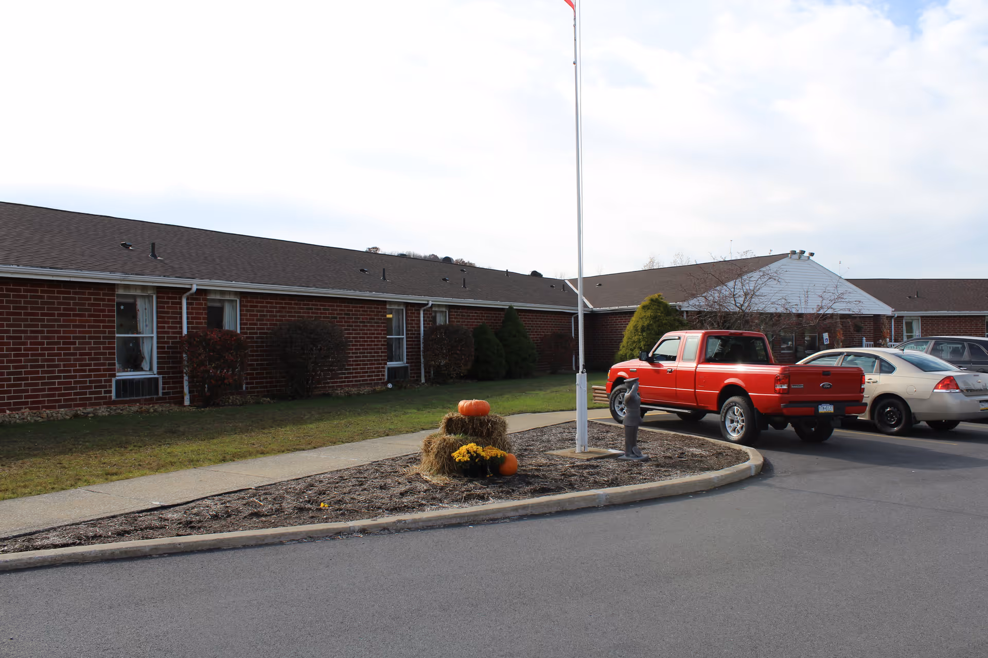 Exterior view of a single-story brick building with a brown roof, a flagpole in the center of a circular landscaped area with hay bales, pumpkins, and yellow flowers. Several cars, including a red pickup truck, are parked along the driveway in front of the building.