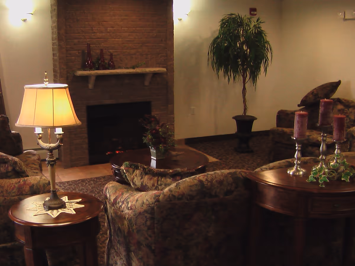 A cozy living room with floral upholstered armchairs and sofas arranged around a wooden coffee table and a fireplace with a brick surround. A table lamp with a beige shade sits on a round wooden side table, and a tall potted plant is placed against the wall. Decorative candles and greenery are on a wooden console table.