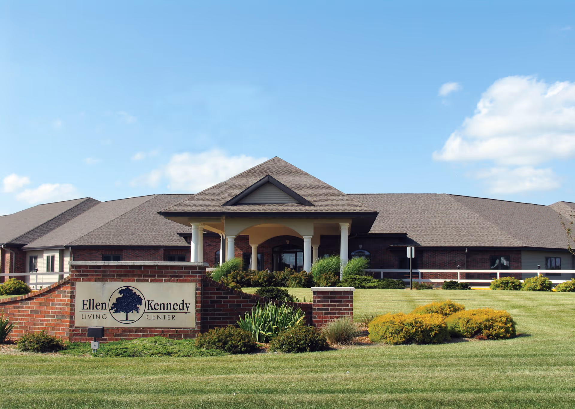 Exterior view of Ellen Kennedy Living Center, a single-story brick building with a covered entrance supported by white columns, surrounded by well-maintained green lawns and shrubs under a blue sky with some clouds.