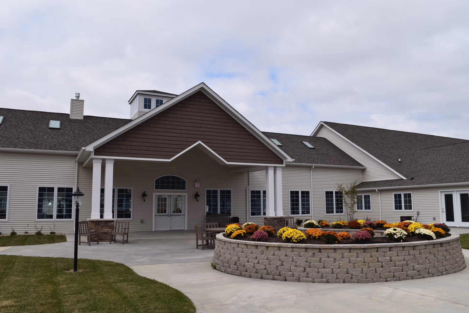 Exterior view of Cedarhurst Senior Living of Jacksonville showing the entrance with a covered porch supported by white columns. There is a circular raised flower bed with colorful flowers in front of the entrance, surrounded by a concrete walkway and green lawn. The building has beige siding and a dark shingled roof under a cloudy sky.