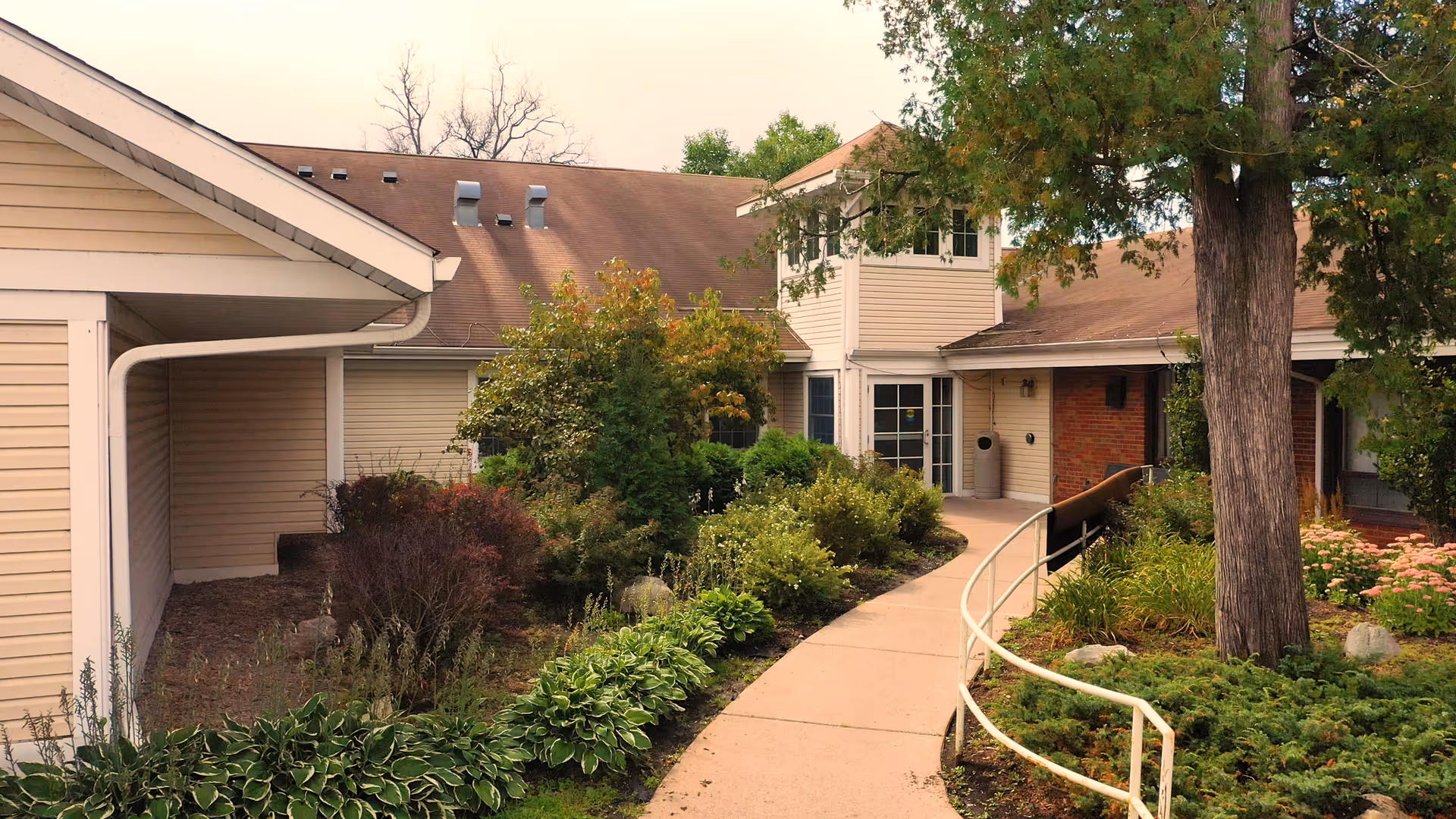 Curved paved walkway and ramp leading to the front entrance of a beige-siding building surrounded by shrubs and trees.