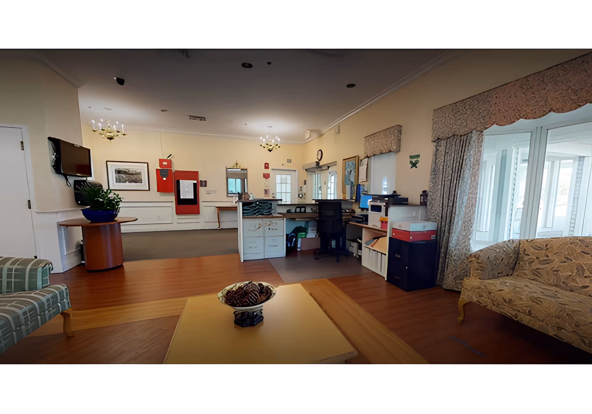 Sunlit reception/lounge area with a front desk, chairs and sofas, a coffee table with a decorative bowl, and large windows with drapes.