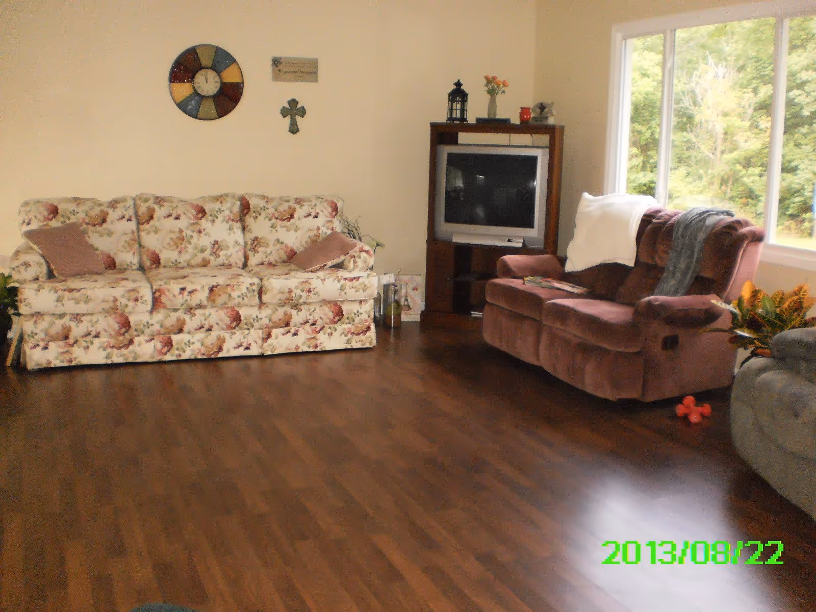 Living room with a floral sofa and brown recliner facing a TV cabinet beside a large window over wood flooring.