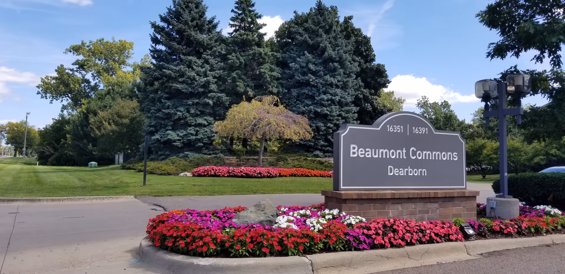 Entrance sign for Beaumont Commons Dearborn set on a brick base surrounded by colorful flowerbeds, trees, and a driveway.
