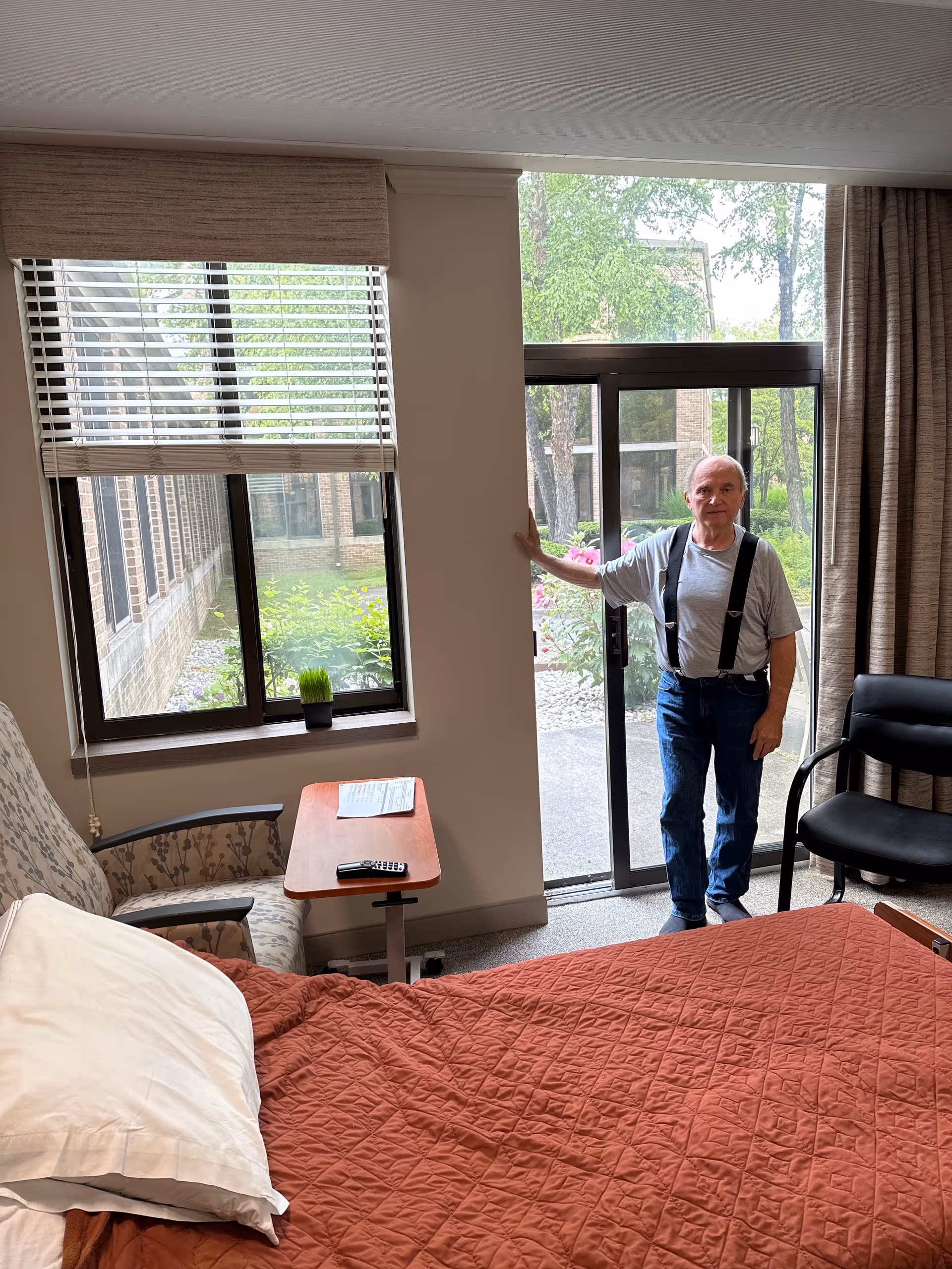 Senior man stands near a glass door in a furnished bedroom with a red quilt bed