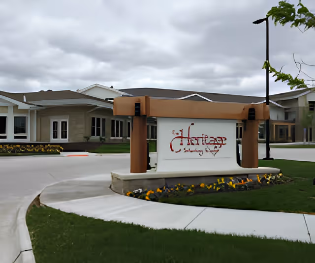 Entrance sign for The Heritage at Sterling Ridge senior living facility with a landscaped flower bed in front and the building visible in the background under a cloudy sky.