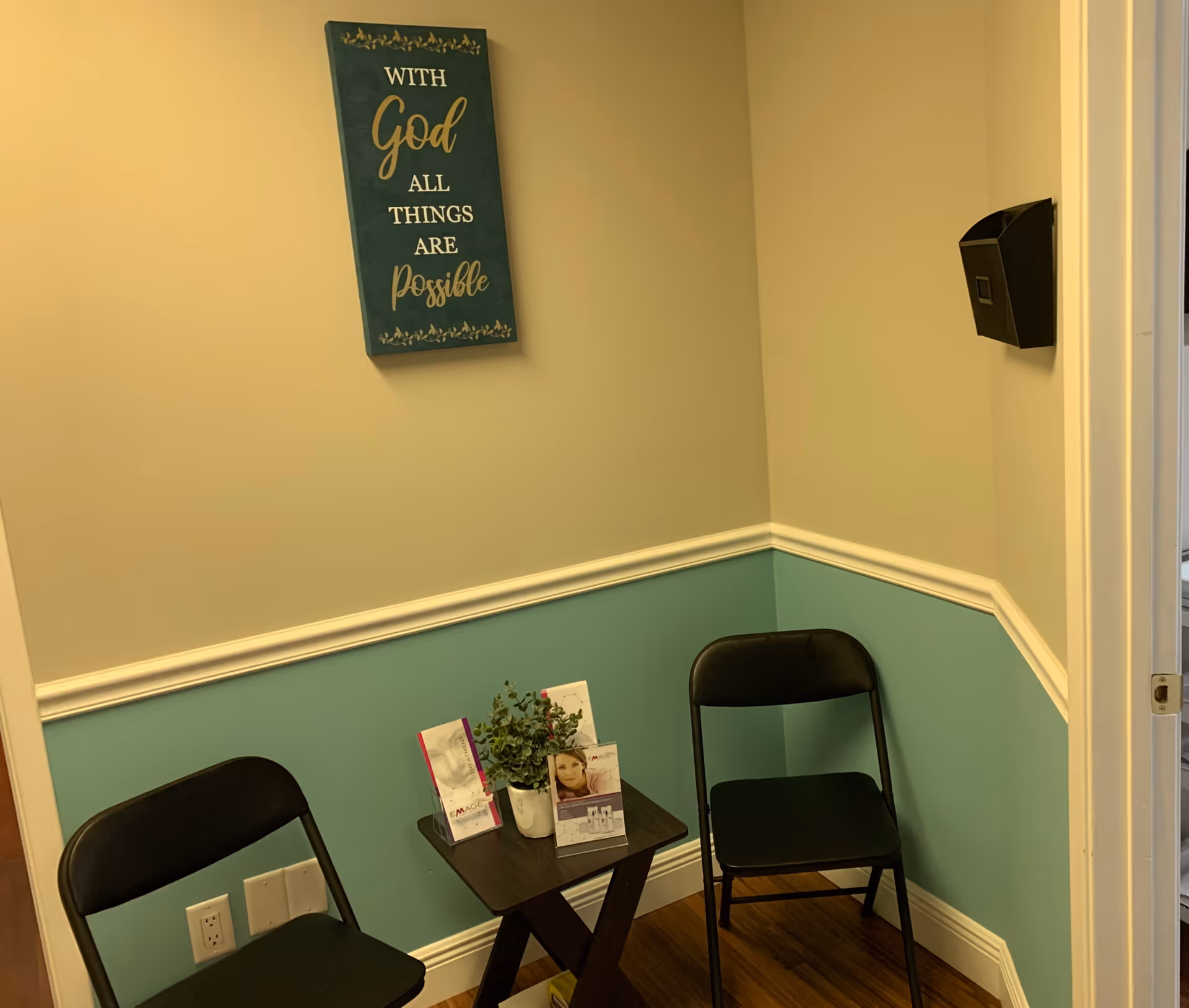 Corner seating area with two folding chairs, a table, plant, and inspirational wall sign.