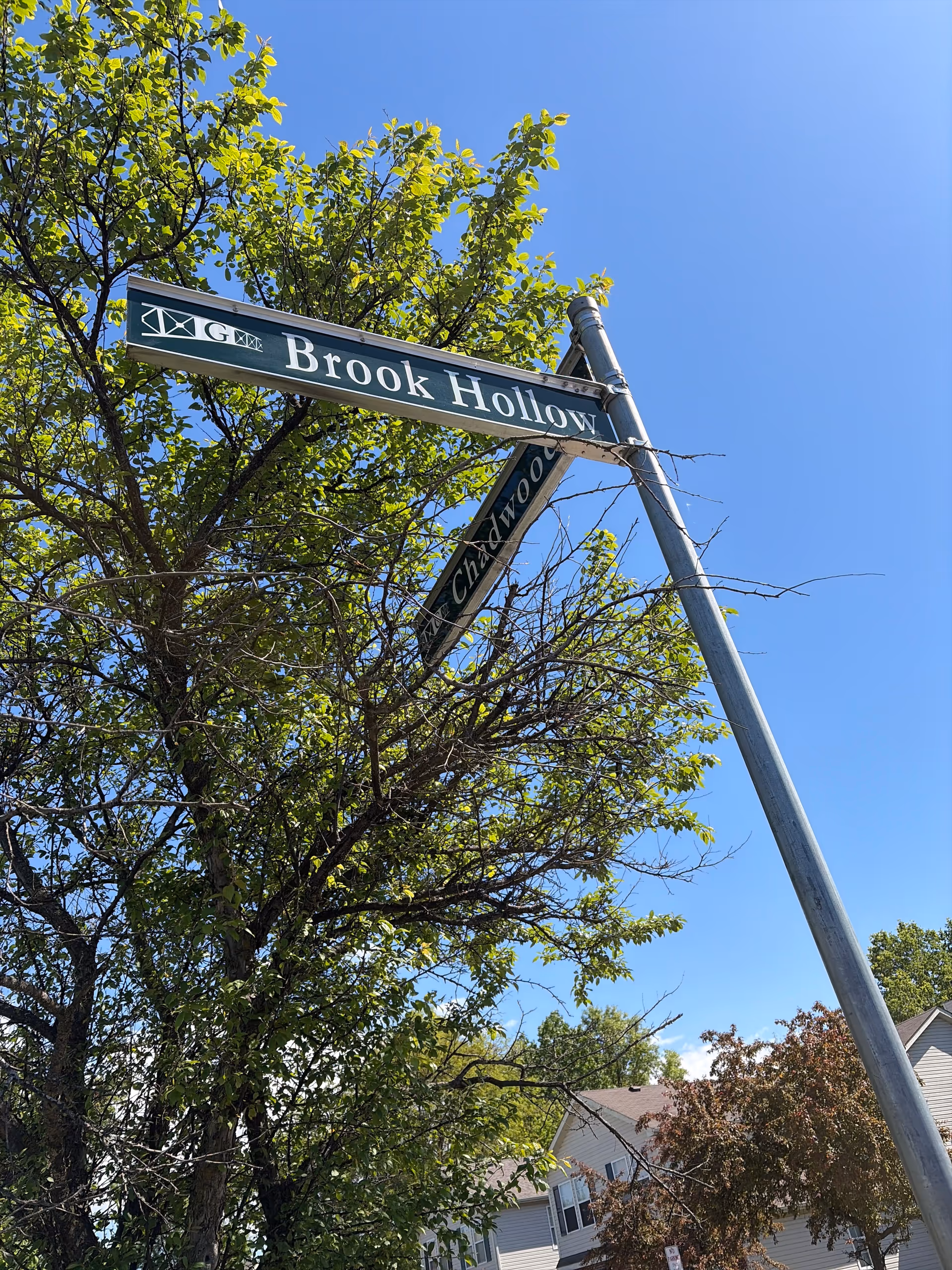 Blue sky above a street intersection with Brook Hollow sign and leafy trees.