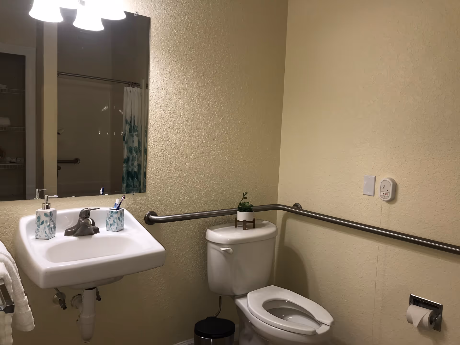 A bathroom with a white sink and faucet, a mirror above the sink, a white toilet with a small potted plant on the tank, a metal grab bar along the wall behind the toilet and sink, a towel hanging on a rack, and a shower curtain visible in the mirror reflection.