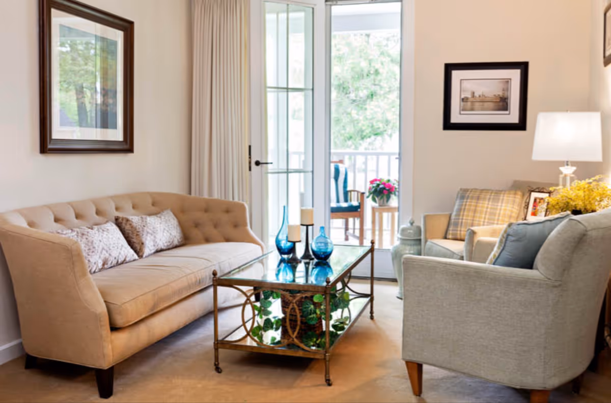 A cozy living room with a beige tufted sofa adorned with patterned pillows, a glass coffee table with blue decorative vases and candles, and two armchairs with plaid and solid cushions. A lamp and framed pictures decorate the walls, and a glass door opens to a porch with a chair and potted flowers.