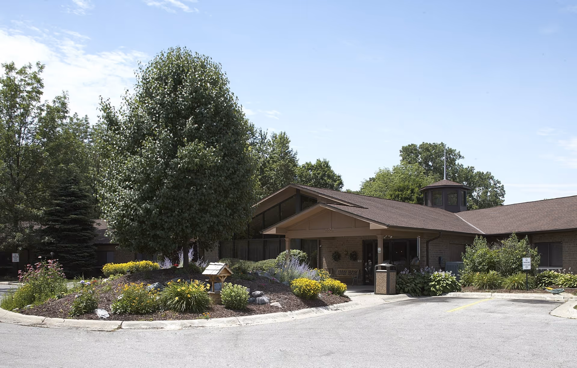 Single-story brick facility entrance with a landscaped circular flower bed, trees, and a paved driveway.